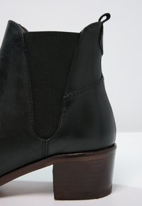 Bottine en cuir noir avec panneau latéral élastique, languette à l'arrière et talon bloc en bois empilé sur fond blanc.