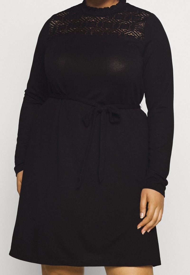 Femme portant une robe noire à manches longues avec empiècement en dentelle et ceinture nouée à la taille, debout devant un fond uni clair.