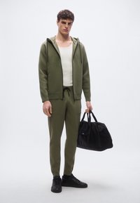 Chándal de punto verde oliva con textura acanalada, sudadera con capucha y cremallera, pantalones joggers a juego y bolso de deporte negro sostenido en una mano. Zapatillas negras.