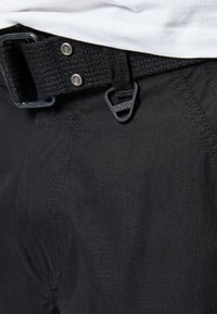 Schwarze Hose mit einem gewebten Gürtel, ausgestattet mit einer metallischen Schnalle und Knopfverschluss. Der Stoff hat eine glatte Textur und einen dezenten Glanz.