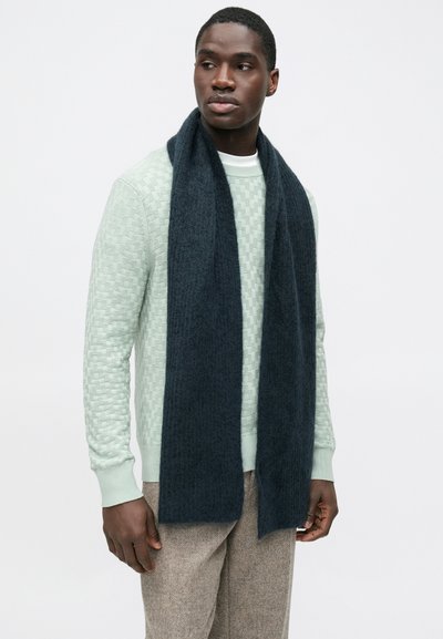 Écharpe bleu foncé en tissu doux, forme rectangulaire allongée, associée à un pull vert menthe texturé, portée par une personne.