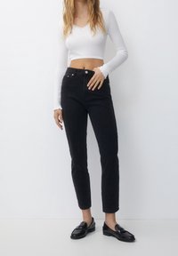 Jean noir taille haute avec jambe fuselée, associé à un haut blanc à manches longues en maille côtelée avec un col et des poignets festonnés.