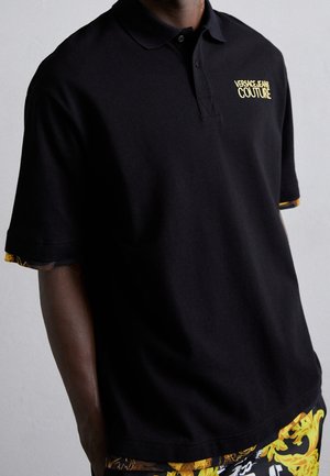 Man met een zwarte Versace Jeans Couture polo met geel logo over een geel-zwart patroon, voor een effen achtergrond.