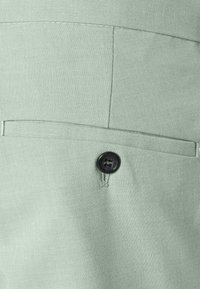 Tuvu skatā gaismas zaļš audums ar melnu pogu, kas aizver horizontālu kabatas atloku uz apģērba.