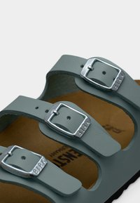 Κοντινό πλάνο γκρι λουριών σανδαλιών με ασημένιες αγκράφες με την επιγραφή "BIRK" πάνω σε καφέ πέλμα, με μερικά ορατά γράμματα.