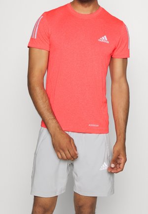 Mann trägt ein hellkorallenrotes Adidas-Shirt mit kurzen Ärmeln und hellgraue Adidas-Shorts, steht mit den Händen nahe der Taille.