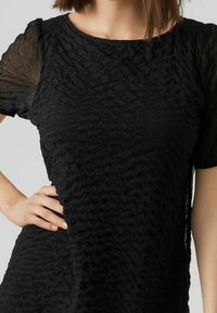 Haut noir texturé avec des manches transparentes courtes, un col rond et un motif ondulé sur le tissu. Drapé subtil sur les côtés.