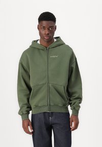 Grüne Freizeitjacke mit Reißverschluss aus Baumwollmischung. Sie verfügt über eine verstellbare Kapuze, Fronttaschen, gerippte Bündchen und ein Logo auf der linken Brustseite.