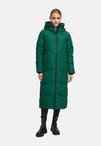 Woman wearing a long, green padded coat with a hood, black pants, and black chunky boots, standing against a plain background.