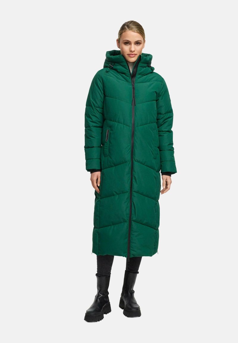 Woman wearing a long, green padded coat with a hood, black pants, and black chunky boots, standing against a plain background.