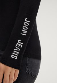 Czarny sweter z długimi rękawami, z białym napisem "JOOP! JEANS" na lewym rękawie. Miękka tekstura i dopasowany krój.