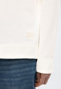 Λευκό μακρυμάνικο πουκάμισο με υφή και ριμπ σχέδιο, με μικρή ουδέτερη ετικέτα στο τελείωμα, σε συνδυασμό με μπλε τζιν παντελόνι.