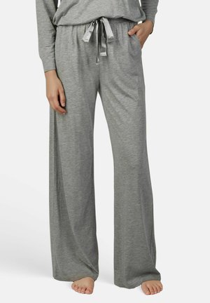Femme portant un pantalon de détente gris ample avec un cordon en satin, debout pieds nus, une main dans la poche et l'autre le long du corps.