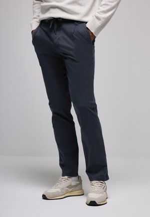 Personne portant un pantalon décontracté bleu foncé et des baskets beiges avec des chaussettes blanches, les mains dans les poches, se tenant devant un fond clair.