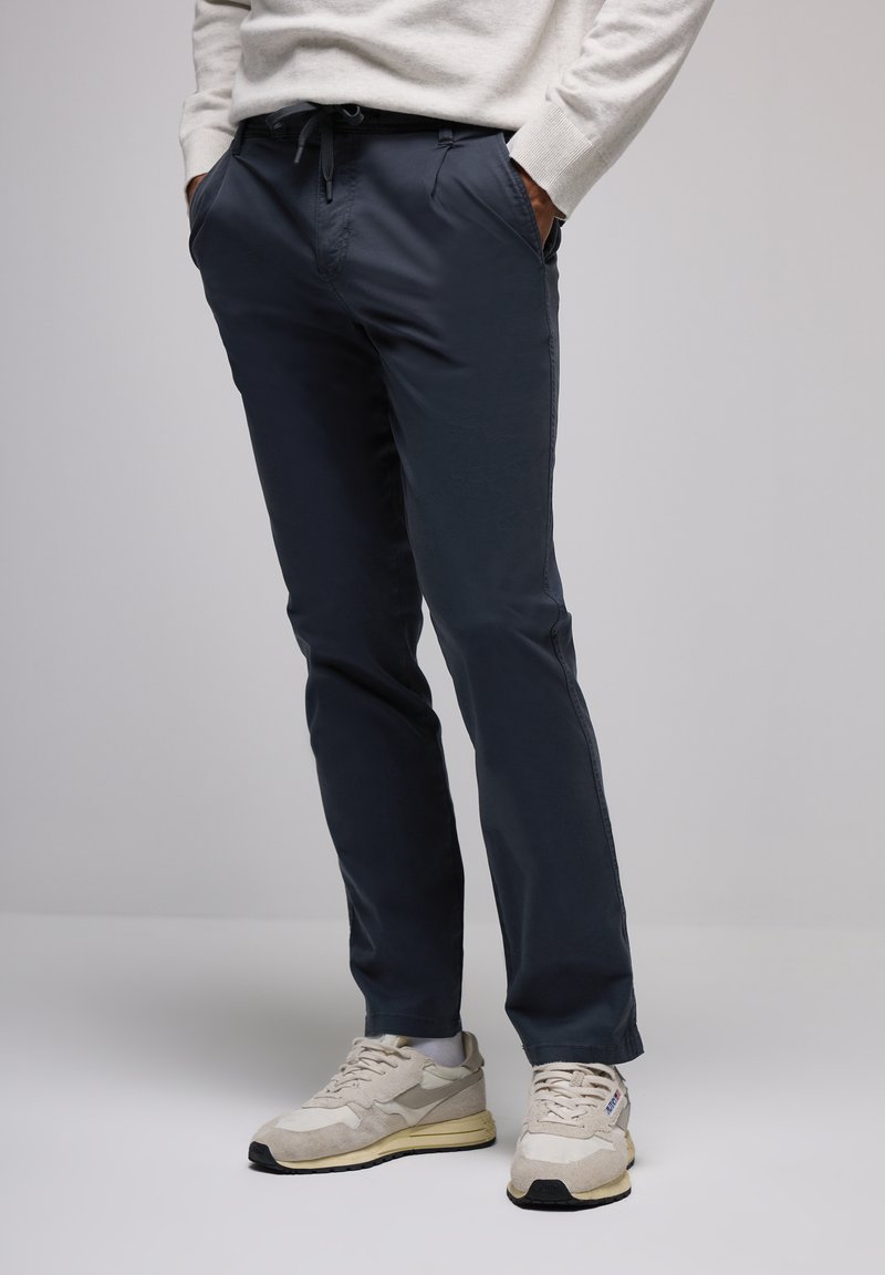 Persona con pantalones casuales azul oscuro y zapatillas beige con calcetines blancos, con las manos en los bolsillos, de pie contra un fondo claro y liso.