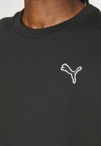 Gros plan d'une personne portant un t-shirt noir à col rond avec un logo Puma contour blanc sur le côté gauche de la poitrine.