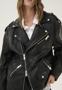 Giacca in pelle nera da motociclista con una finitura testurizzata, cerniere in argento e diverse tasche con cerniera. Presenta spalline sulle spalle e un ampio colletto.