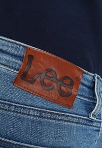 Denim jeans featuring a brown leather patch with the engraved "Lee" logo, blue denim fabric, light wash with visible stitching details.