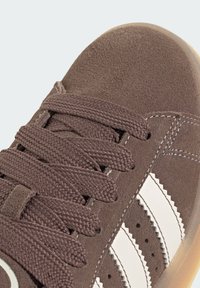 Brown suede sneaker with flat brown laces, white striped accents, and a textured rubber sole. Features perforated details on the side.