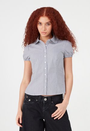 Giovane donna con capelli ricci e rossi che indossa una camicia a righe a maniche corte con bottoni e pantaloni neri, in piedi davanti a uno sfondo semplice.