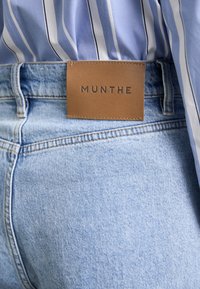 Lyseblå denimjeans med et brunt lædermærke, der viser "MUNTHE." Jeansene har subtile falmninger og standard syninger.