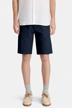 Homme portant un short bleu marine, une chemise à boutons blanc cassé, des chaussettes gris clair et des chaussures décontractées beige, debout devant un fond uni.