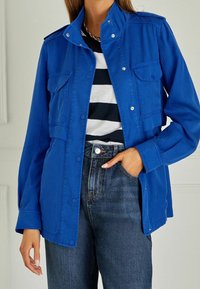 Chaqueta de mezclilla azul con botones a presión, dos bolsillos frontales y un cuello alzado, combinada con una camisa a rayas negras y blancas y jeans oscuros.