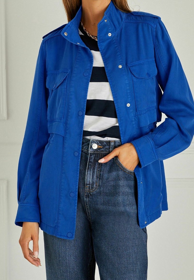 Chaqueta de mezclilla azul con botones a presión, dos bolsillos frontales y un cuello alzado, combinada con una camisa a rayas negras y blancas y jeans oscuros.