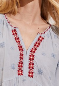 Błękitna bluzka z czerwonym haftem kwiatowym wokół dekoltu i rękawów. Materiał wydaje się miękki i lekki, a talia jest marszczona.