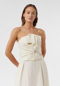 Beżowy top bez ramiączek z twisted front, wykonany z gładkiego materiału o subtelnej fakturze. Brak widocznych elementów metalowych lub wzorów.