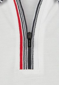 T-shirt polo blanc avec un col zippé, présentant des rayures rouges et noires sur le col. Tissu texturé et détail de fermeture éclair noir.