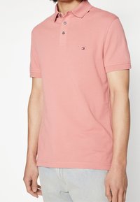 Rosa pikétröja av bomull med krage, två knappar, korta ärmar och en liten logotyp på vänster bröst.