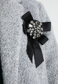 Broche ruban noir avec un cluster de pierres précieuses claires fixé sur un vêtement en tissu gris texturé.