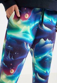 Kolorowe spodnie dresowe z żywym nadrukiem galaktyki w odcieniach niebieskiego, zielonego i fioletowego. Zawierają motywy gór i elementy niebiańskie. Miękka tkanina.