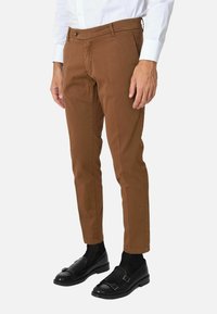 Pantaloni slim-fit marroni in cotone, con chiusura frontale a bottone, tasche laterali e una texture liscia. Indossati con mocassini neri.