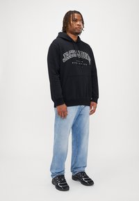 Sort hættetrøje med "JACK & JONES" trykt på brystet, parret med lyseblå jeans og sorte sneakers med lilla detaljer.