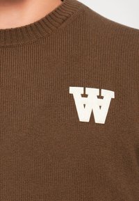 Hnedý rebrovaný sveter s okrúhlym výstrihom, na ľavej strane hrude so štruktúrovaným bielym logom "W".