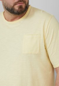 Een bebaarde man die een lichtgele T-shirt met korte mouwen en een borstzak draagt, tegen een effen achtergrond.