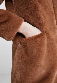 Brown faux fur coat with a soft texture and side pockets. Features a relaxed fit and slightly oversized sleeves. Close-up view of hand in pocket.