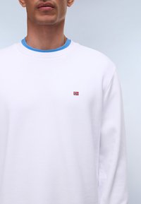 Λευκό βαμβακερό φούτερ με μακριά μανίκια, στρογγυλή λαιμόκοψη και μικρό κεντητό λογότυπο με σημαία στο στήθος. Η εσωτερική λαιμόκοψη έχει μπλε λεπτομέρεια.