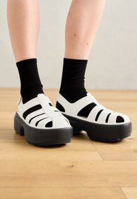 White rubber sandals with black accents, featuring open toes and cut-out sides. Chunky gray soles provide height and textured grip.