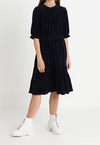 Abito blu navy con un orlo arricciato, maniche a sbuffo e vita arricciata, abbinato a stivaletti bianchi con lacci sopra la caviglia.