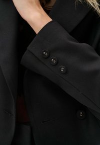 Czarna marynarka o zwartej konstrukcji, miękkim materiale i trzech czarnych guzikach na mankietach z metalowymi pętlami. Dopasowany fason.