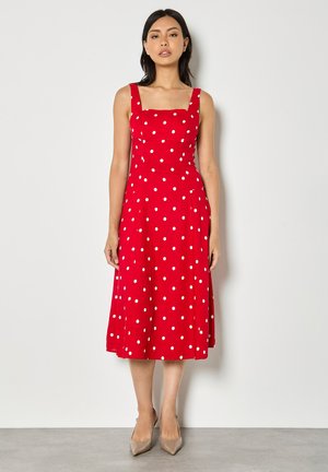 Kobieta stojąca, ubrana w czerwoną, bezrękawnikową midi sukienkę w białe kropki, oraz beżowe buty na obcasie z ostrym noskiem, na tle białej ściany.
