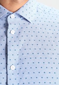 Camisa de algodón azul claro con un patrón de pequeñas flores azul oscuro, con un cuello y botones blancos en la parte delantera.