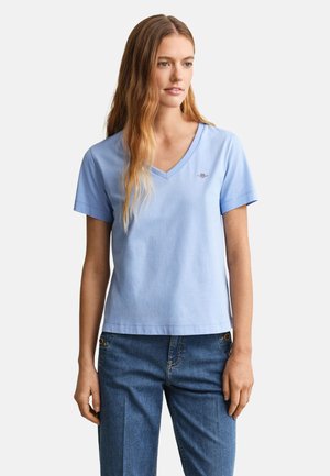 Fiatal nő világoskék V-nyakú, rövid ujjú pólóban és kék farmerben, semleges arckifejezéssel áll egy egyszerű háttér előtt.