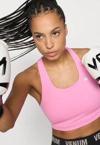 Ružová športová podprsenka s zaobleným výstrihom, hladkou textúrou a jemným logom. Čierne šortky majú pás Venum so zlatými akcentmi.