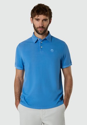 Homme aux cheveux bruns courts et à la barbe, portant un polo bleu à manches courtes et un pantalon de couleur claire, sur un fond uni.
