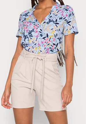 Woman wearing light blue short-sleeve blouse with multicolor floral print and high-waisted beige shorts with tie belt.
