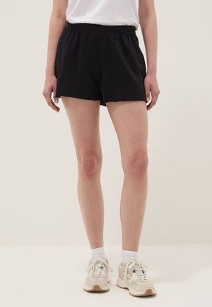 Persoon die zwarte shorts met elastische taille, witte sokken en beige sneakers draagt, staand tegen een effen lichte achtergrond.
